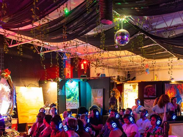 Crowd at a Lemonade Arts gallery activation — projections, paintings, disco ball, and audience wearing illuminated silent-disco headphones.