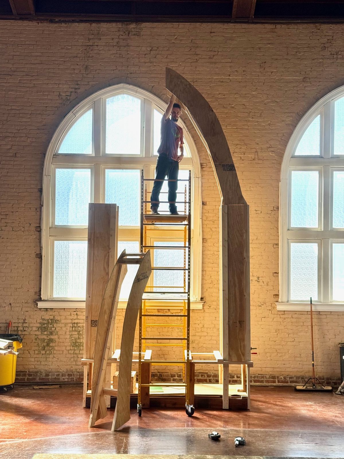 An artist on scaffolding installs a sculptural arch inside a historical brick building with arched cathedral windows in St. Petersburg.